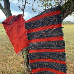 May include: Two woven rugs, one solid red and the other with red stripes on a dark grey background, are draped over a tree branch. The rugs have fringed edges and a rustic, handmade appearance. The scene is set outdoors with a cloudy sky.