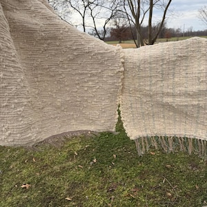 May include: A beige woven textile with a textured surface and fringe detail. The textile is hanging outdoors, with a green grassy background and bare trees in the distance. The fabric appears to be thick and warm.