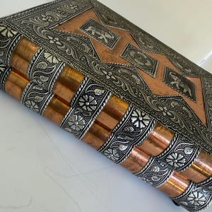 May include: An ornate, rectangular box with a copper-colored exterior and silver-colored filigree detailing. The box features floral and geometric patterns, with a central design of elephants. The edges are adorned with a repeating floral motif.