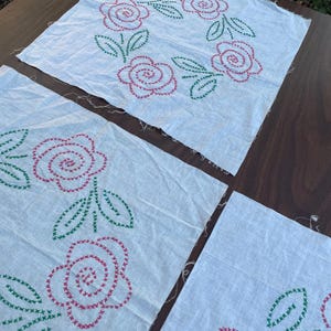 May include: Four white fabric squares with hand-stitched floral designs. The flowers are red, and the leaves are green. The stitching is visible, and the fabric has a slightly wrinkled texture. The squares are arranged on a brown surface.