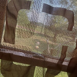 May include: A sheer brown curtain with a repeating horse design. The curtain is made of a woven fabric, allowing light to filter through. The design is visible against a backdrop of green foliage.