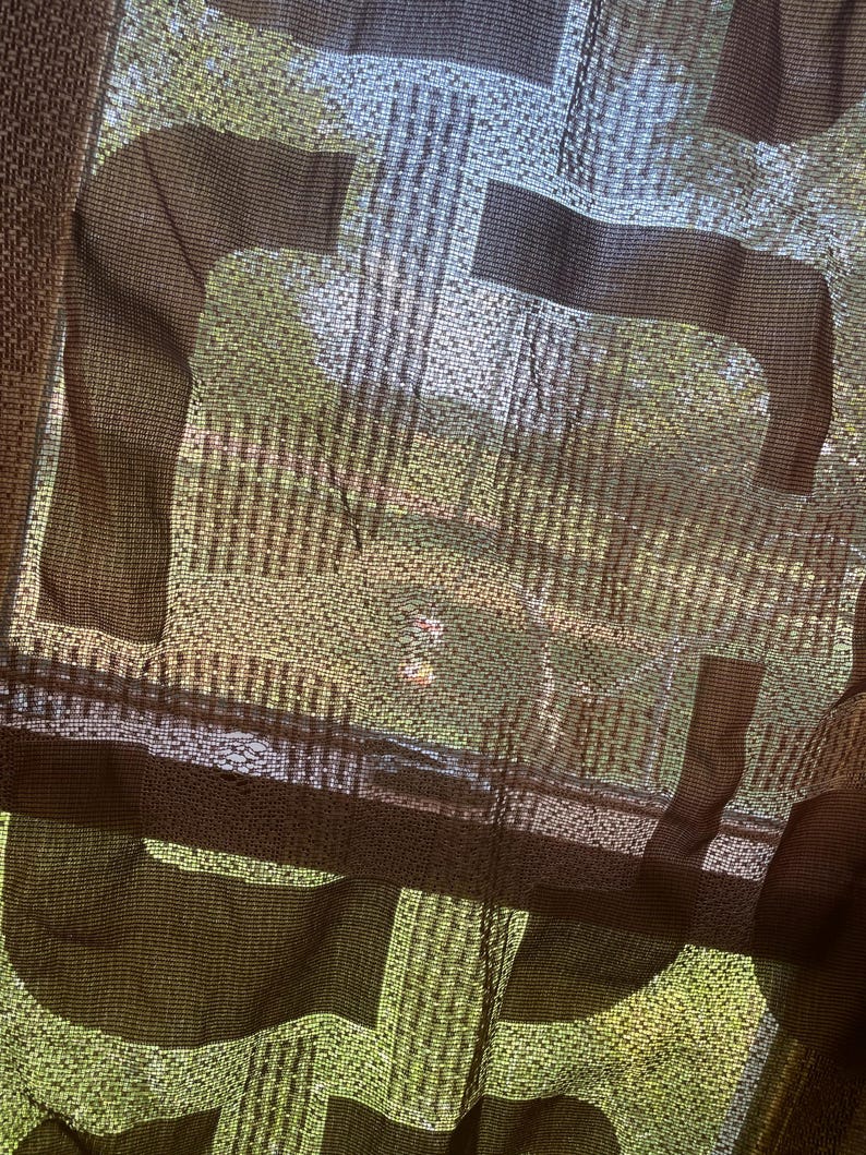 May include: A sheer brown curtain with a repeating horse design. The curtain is made of a woven fabric, allowing light to filter through. The design is visible against a backdrop of green foliage.