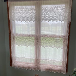 May include: White lace curtains with scalloped edges hang in a window. The curtains are layered with three tiers of lace.