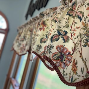 May include: Cream-colored floral valance with a repeating pattern of flowers and leaves in shades of red, blue, and green. The valance is trimmed with a dark red decorative border and is hanging in front of a window.