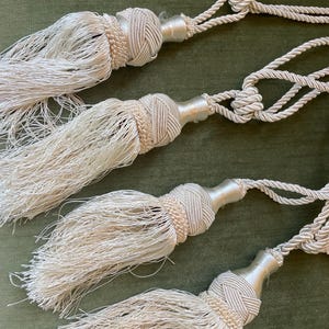 May include: Four white tassel tiebacks with a braided rope design. The tassels are made of a soft, fluffy material and have a beaded detail.