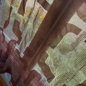 May include: Close-up of a sheer curtain with a geometric pattern. The curtain features a repeating design of brown and white shapes against a light green background. The fabric is translucent, allowing light to filter through.