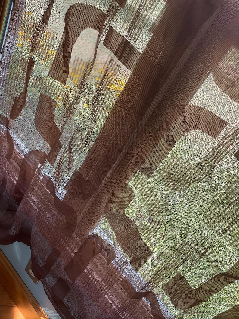 May include: Close-up of a sheer curtain with a geometric pattern. The curtain features a repeating design of brown and white shapes against a light green background. The fabric is translucent, allowing light to filter through.