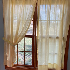 May include: A pair of yellow gingham curtains with a white background hang in a window. The curtains are tied back on each side, revealing a view of a yard with trees and grass.