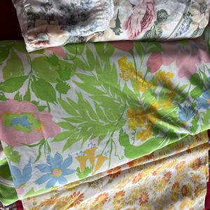 May include: Three different floral patterned fabric swatches. The top swatch is a white fabric with a pink and blue floral pattern. The middle swatch is a white fabric with a green, pink, yellow, and blue floral pattern. The bottom swatch is a white fabric with a yellow and orange floral pattern.