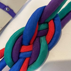 May include: A close-up of a colorful belt with a woven design. The belt features a braided pattern with strands of red, blue, green, and purple fabric. A portion of a beige belt strap with a purple loop is visible.