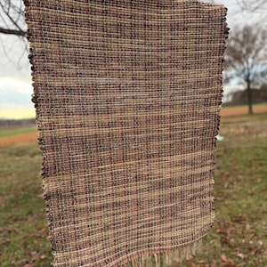 May include: A rectangular woven rug with a plaid pattern in shades of brown, tan, and burgundy. The rug has a fringed edge and is hanging from a tree branch outdoors. The rug appears to be made of natural fibers.