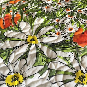 May include: A quilted fabric featuring a vibrant floral pattern with large white daisies, bright orange poppies, and green foliage. The design includes black line work details and yellow centers on the daisies, all set against a white background.