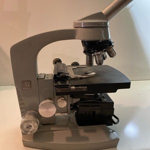 May include: A gray vintage microscope with a black stage and a silver knob. The microscope has a black and silver objective lens turret and a silver focusing knob.