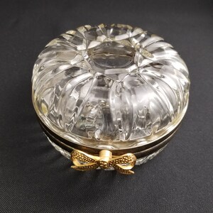 May include: A clear, round glass trinket box with a ribbed design. The box has a gold-colored bow clasp and a dark metal band around the middle. The lid has a circular opening in the center.