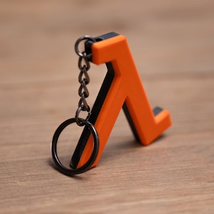 May include: A bright orange and black Lambda symbol keyring. The keyring has a black key ring and chain. The Lambda symbol is a stylized letter, with a black outline and a bright orange interior. It rests on a wooden surface.