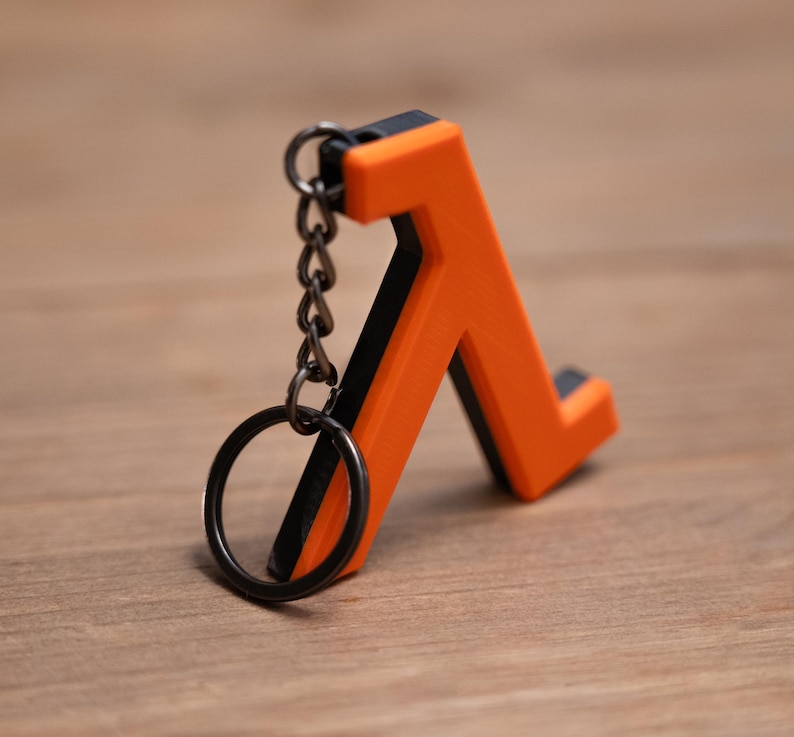 May include: A bright orange and black Lambda symbol keyring. The keyring has a black key ring and chain. The Lambda symbol is a stylized letter, with a black outline and a bright orange interior. It rests on a wooden surface.