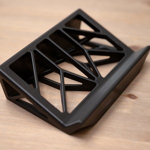 May include: A black, geometric, 3D printed stand for a phone or tablet. The stand has a triangular base and a raised platform with an open, lattice-like design.