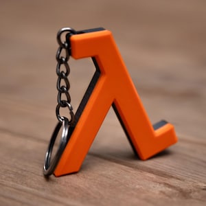 Peut inclure: Un porte-clés symbole Lambda orange et noir. Le porte-clés présente un symbole Lambda orange vif avec un bord noir, attaché à une chaîne et un porte-clés argentés. Le porte-clés est en matériau solide, posé sur une surface en bois.