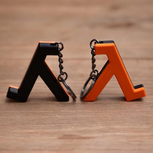 May include: Two keyrings, one black and one orange, each with a stylized letter "A" design. The keyrings have a metal chain and ring attachment. The background is a light brown wooden surface.