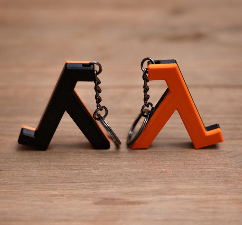 May include: Two keyrings, one black and one orange, each with a stylized letter "A" design. The keyrings have a metal chain and ring attachment. The background is a light brown wooden surface.