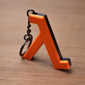 May include: A two-tone keychain featuring an orange and black lambda symbol. The keychain has a black chain and ring. The lambda symbol is a stylized letter, commonly used in mathematics and physics.