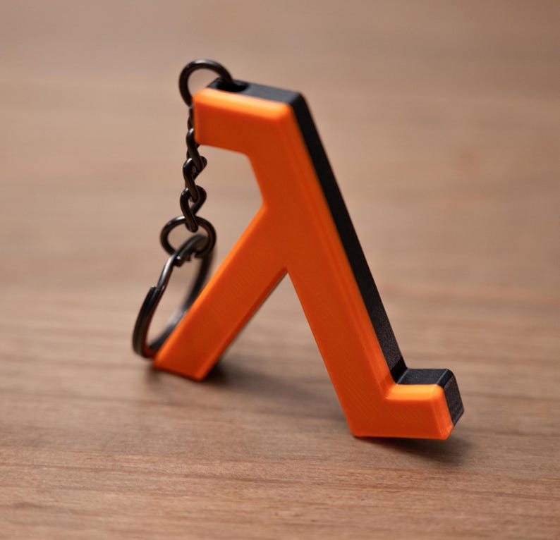 May include: A two-tone keychain featuring an orange and black lambda symbol. The keychain has a black chain and ring. The lambda symbol is a stylized letter, commonly used in mathematics and physics.