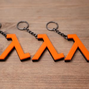 May include: Three orange and black keyrings featuring a lambda symbol design. Each keyring has a silver-coloured chain and ring. The keyrings are displayed on a wooden surface.