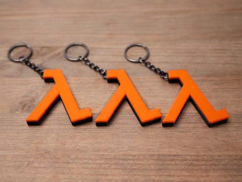 May include: Three orange and black keyrings featuring a lambda symbol design. Each keyring has a silver-coloured chain and ring. The keyrings are displayed on a wooden surface.