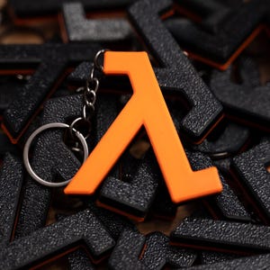 May include: An orange lambda symbol keyring with a silver ring. The lambda symbol is prominent, set against a backdrop of many black lambda keyrings. These keyrings are a gaming accessory.