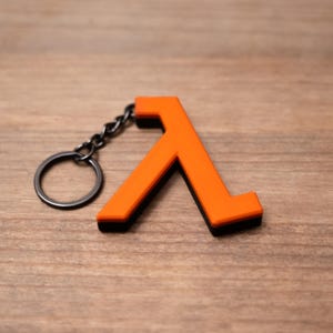 May include: An orange Lambda symbol keyring with a black border and a silver key ring. The Lambda symbol is a stylized letter used in the Half-Life video game series. The keyring is made of a durable material and is designed to attach to keys or a bag.