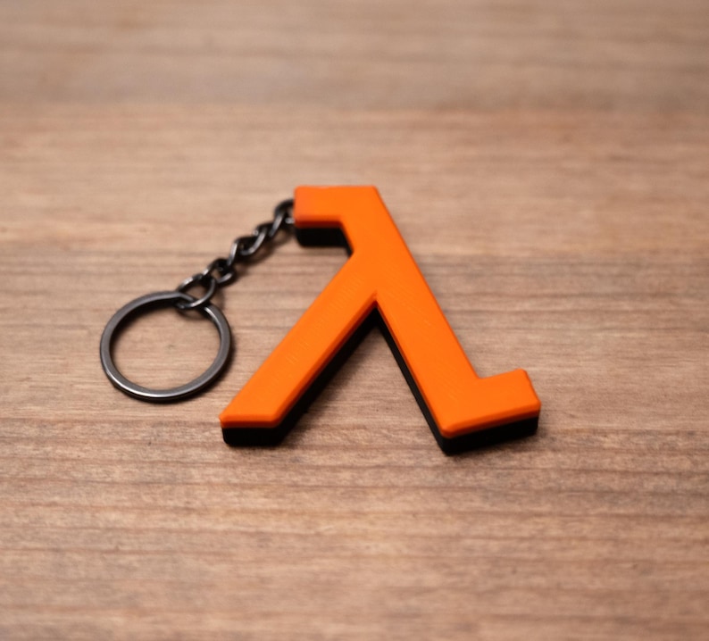 May include: An orange Lambda symbol keyring with a black border and a silver key ring. The Lambda symbol is a stylized letter used in the Half-Life video game series. The keyring is made of a durable material and is designed to attach to keys or a bag.