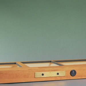 May include: A rectangular wooden display case with three recessed compartments. The case is made of light-colored wood and features a brass plate with two holes and a power outlet. The word "ZEPHYR" is etched on the side.