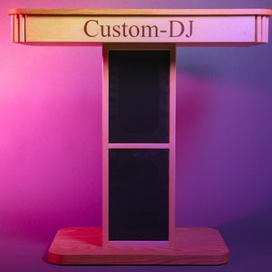 May include: A wooden DJ stand with the text "Custom-DJ" on the top. The stand has a T-shape design with a rectangular base and a top. The center section features a black mesh panel. The stand is made of light-coloured wood.