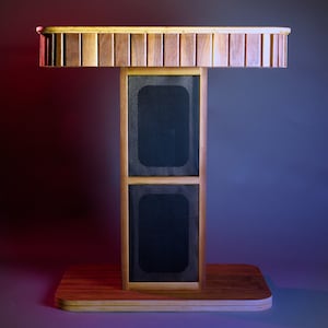 May include: A wooden speaker stand with a rectangular base and a T-shaped structure. The stand features two black speaker covers and a top section with vertical wooden slats. The wood has a natural finish.