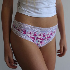 Op de afbeelding: Slip met bloemenprint en witte kanten rand. Het patroon bevat roze, paarse en blauwe bloemen. De taille en beenopeningen zijn afgezet met delicate witte kant.