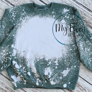 Puede incluir: Una sudadera de color verde azulado con un diseño desteñido y desgastado. La sudadera presenta una gran zona blanca desteñida en la parte delantera y salpicaduras blancas por todas partes. El texto "My Five Designs" está en escritura negra dentro de un círculo verde azulado.