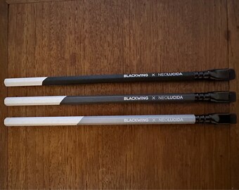 May include: Three black and white pencils with a black eraser and the text "BLACKWING x NEOLUCIDA" printed on each pencil.
