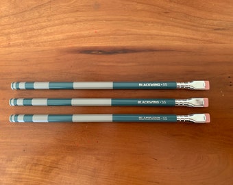 May include: Three teal and silver pencils with black and white striped barrels and pink erasers. The pencils are labeled "Blackwing 55".