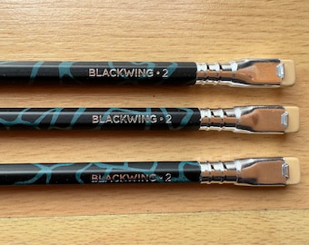 May include: Three black pencils with a blue and black patterned design. Each pencil has a silver metal ferrule and a beige eraser. The pencils are labeled "BLACKWING 2".