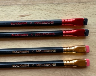 May include: Four black pencils with gold and red accents. The pencils are labeled "BLACKWING X MOLESKINE" and have different hardness grades: "SOFT", "SOFT", "2H", and "HB".