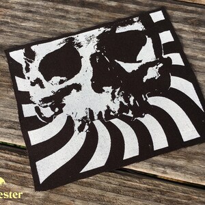 Puede incluir: Parche cuadrado en blanco y negro con un diseño de calavera. La calavera blanca, de aspecto desgastado, se encuentra sobre un fondo de rayas blancas y negras. Ideal para ropa o accesorios.