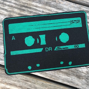 Op de afbeelding: Een zwart rechthoekig embleem met een groen cassettebandontwerp. Het embleem bevat de tekst "A", "DR" en "60", samen met andere tekst. Het ontwerp doet denken aan vintage audiotechnologie.