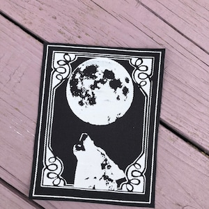 May include: Black and white patch featuring a howling wolf under a full moon. The design is framed with a decorative border. The patch is rectangular and appears to be made of fabric.