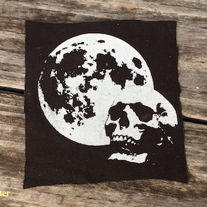 May include: A dark brown square patch featuring a white graphic of a full moon and a skull. The moon is detailed with craters, and the skull is positioned below it. The patch has a raw edge.