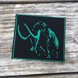 Puede incluir: Un parche cuadrado negro con un borde turquesa y una ilustración turquesa de un mamut lanudo. El mamut está representado en un diseño simple y estilizado. El parche es probablemente de tela y es adecuado para coser o planchar en ropa o accesorios.