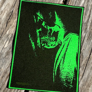 Puede incluir: Parche rectangular negro con una ilustración verde neón de una figura con forma de calavera con una capa con capucha. El diseño está delineado en verde neón. El parche está sobre una superficie de madera desgastada.
