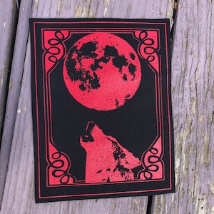 Pode incluir: Remendo retangular preto com design vermelho. O design apresenta uma lua cheia e um lobo uivando, emoldurados por uma borda decorativa vermelha. Adequado para roupas.