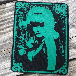 Könnte beinhalten: Ein rechteckiger schwarzer Aufnäher mit einer grünen Silhouette einer Frau mit Sonnenbrille. Das Bild hat eine Vintage-Ästhetik im Siebdruck, mit einem dekorativen grünen Rand. Das Haar der Frau ist im Retro-Stil frisiert.