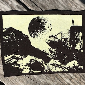 Peut inclure: Un écusson rectangulaire avec une bordure noire et un intérieur crème. L'écusson présente une illustration en noir et blanc d'une personne debout sur une falaise, avec une grande sphère et un paysage montagneux.