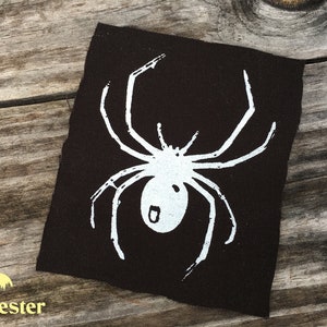 May include: A dark brown fabric patch featuring a white spider illustration. The spider has eight legs and a rounded body. The patch is placed on a wooden surface. The word "Harvester" is printed at the bottom.